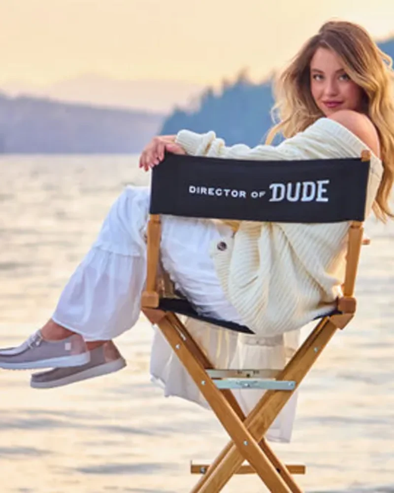 Sydney Sweeney seated in a Director of Dude chair beside a lake at sunset for Hey Dude